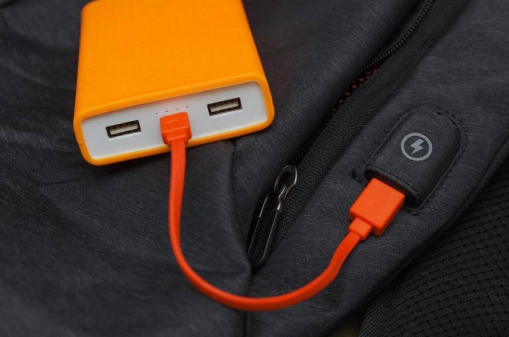 A bright orange power bank with USB ports connected to an orange charging cable, placed on a dark fabric surface.