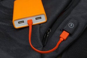 A bright orange power bank with USB ports connected to an orange charging cable, placed on a dark fabric surface.