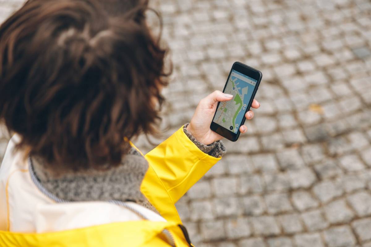 A person in a yellow jacket looks at a smartphone displaying a map while standing on a cobblestone street.