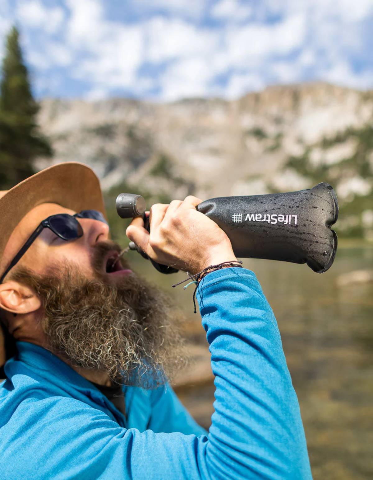A person in a blue jacket drinks from a black water bottle while enjoying a scenic outdoor landscape with mountains and trees in the background.