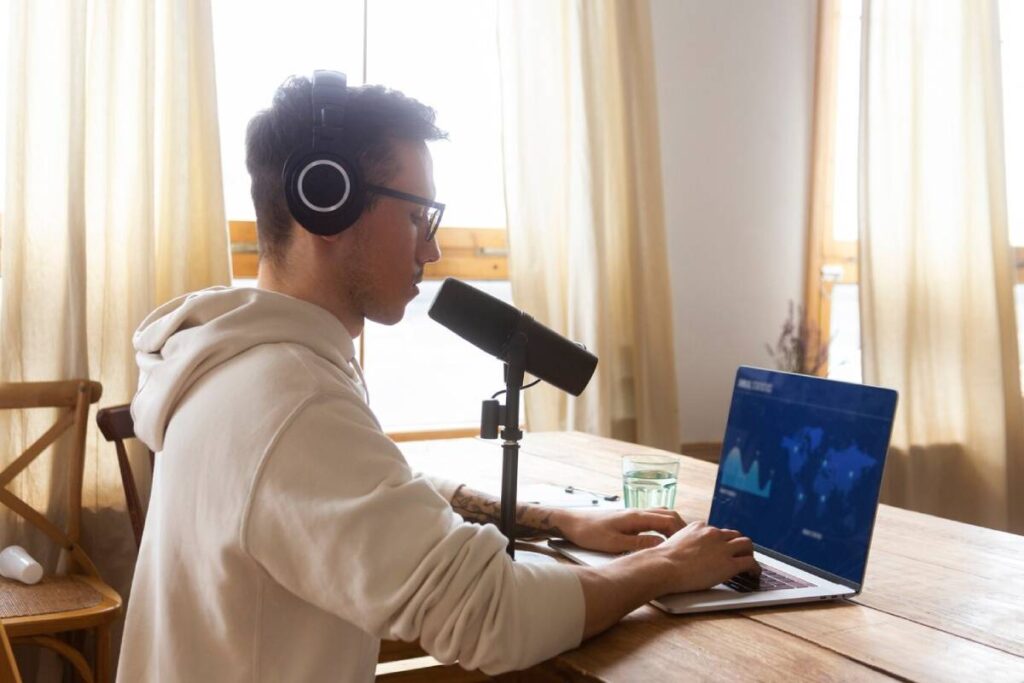 A person wearing headphones works at a laptop with a microphone, set in a bright room with large curtains and a glass of water nearby.
