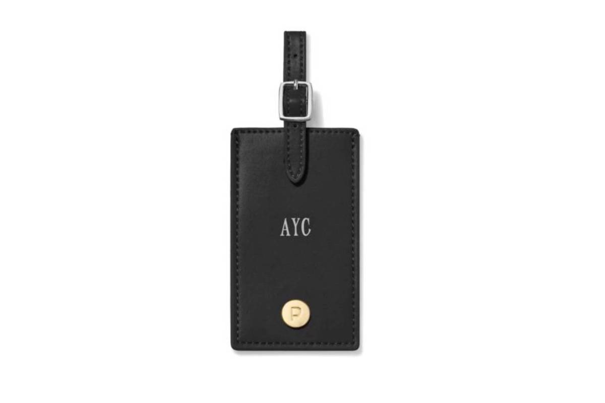 Black leather luggage tag with embossed AYC and a gold P emblem, featuring a strap and silver buckle for attachment.
