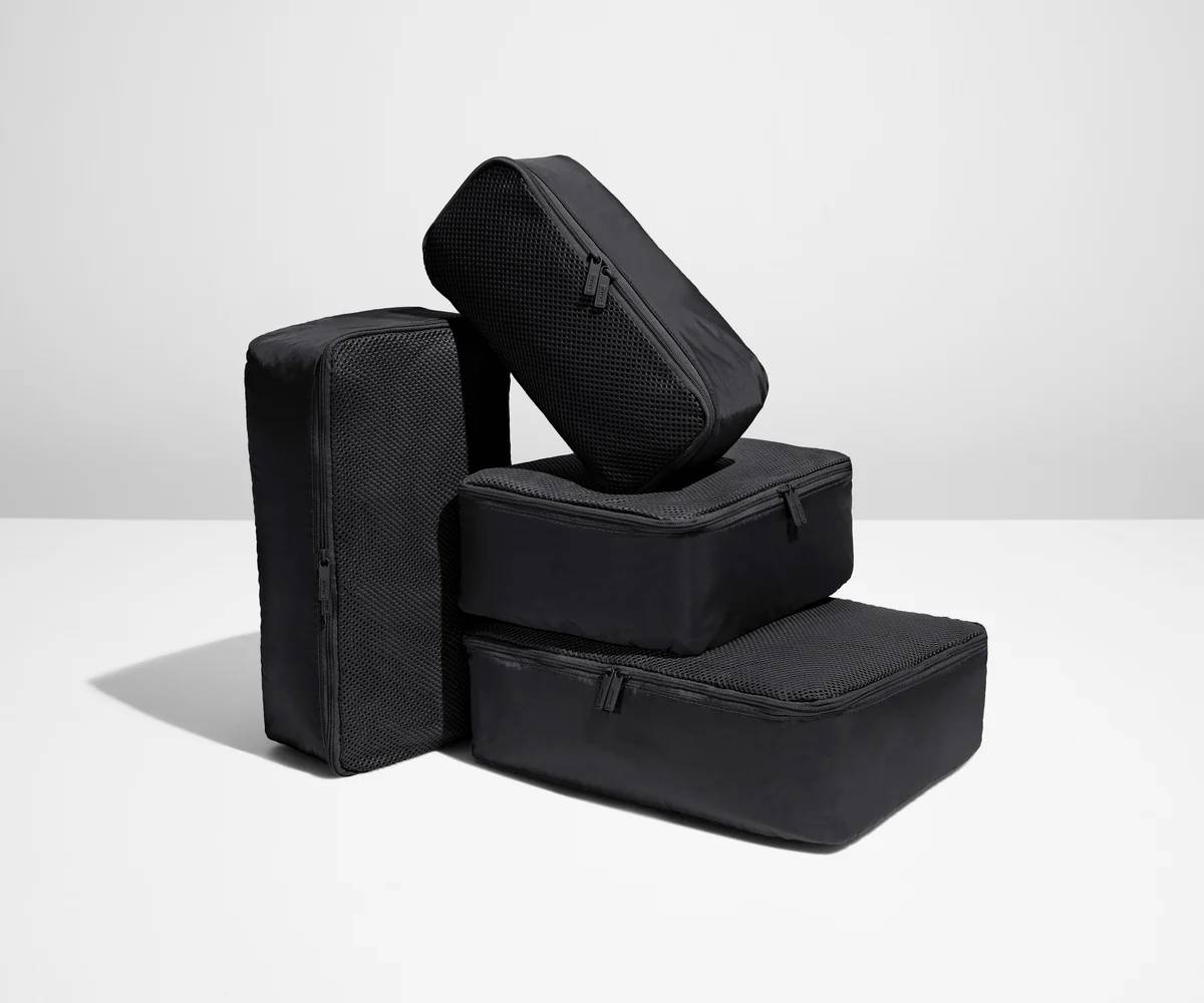 Four black packing cubes of various sizes, stacked and arranged on a white surface against a minimalist background. 