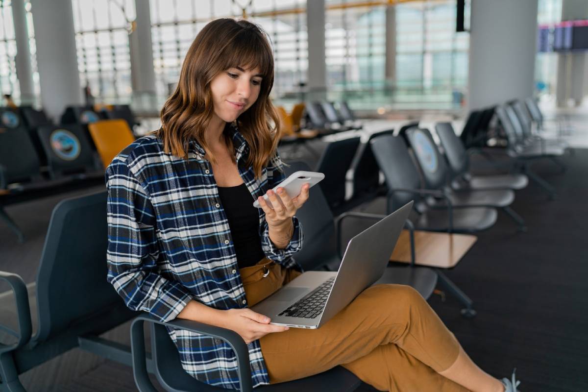  A woman sits in an airport lounge, using a smartphone while a laptop rests on her lap, dressed casually in a flannel shirt and pants.