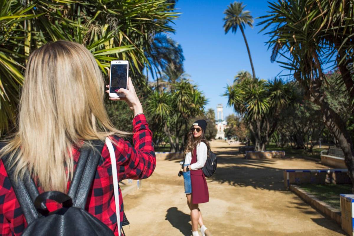 A woman takes a selfie against a backdrop of palm trees and a bright blue sky, while another woman stands in the distance. 