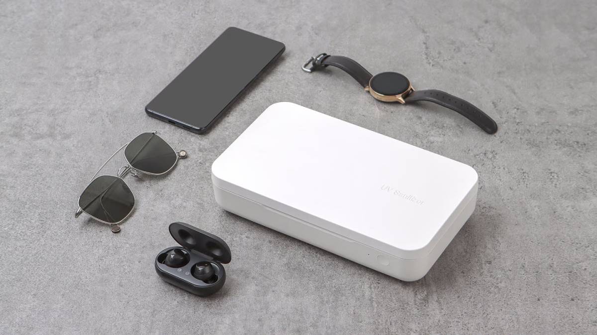 A white UV sanitizer box surrounded by sunglasses, a smartphone, a smartwatch, and wireless earbuds on a gray surface.