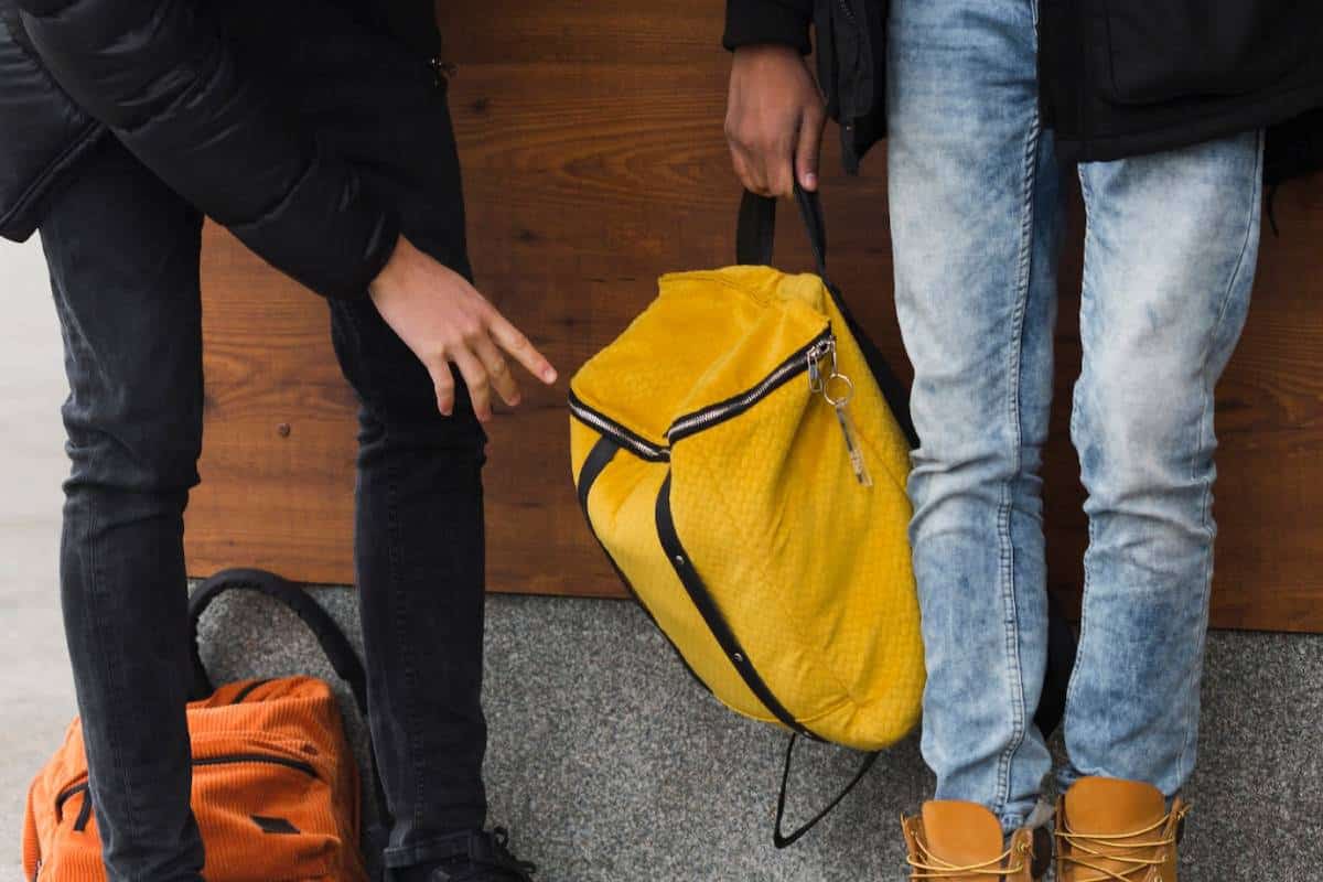 Two individuals stand beside a wooden wall; one points at a bright yellow bag held by the other, who wears denim and boots.