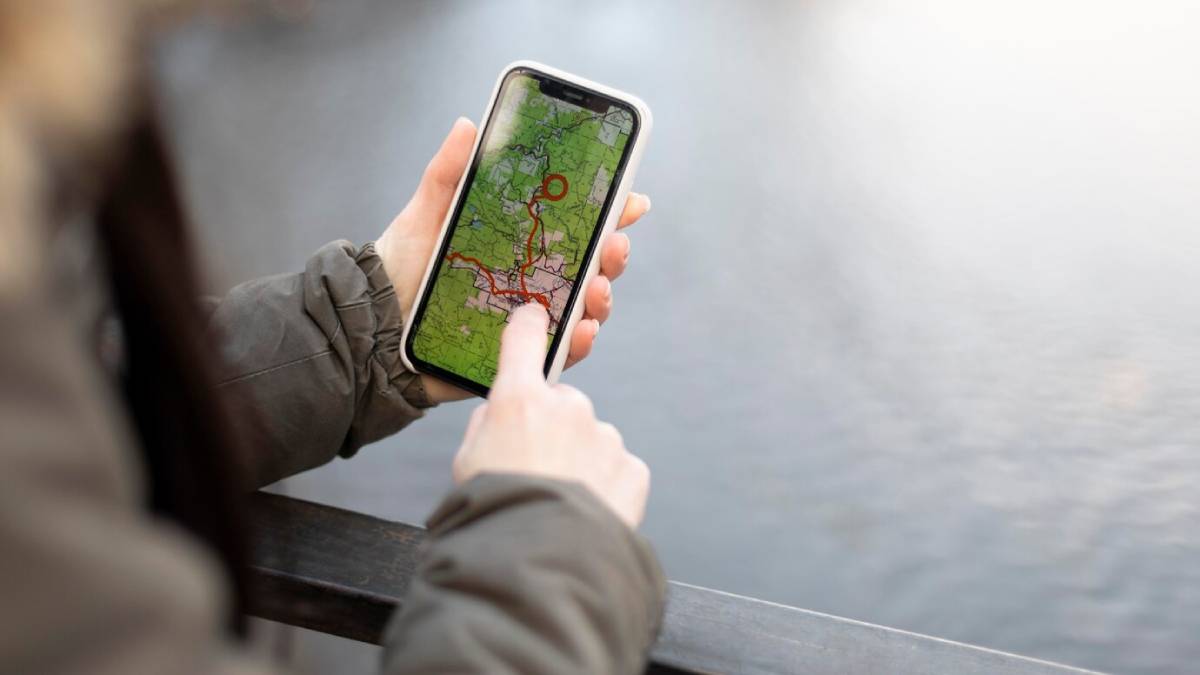 A person holds a smartphone displaying a detailed map with marked routes, while standing by a body of water.