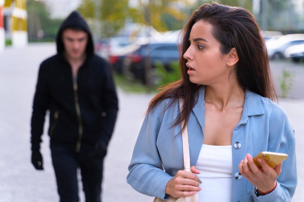 A woman walks down a city sidewalk, holding her phone, while an individual in a black hoodie approaches behind her. 