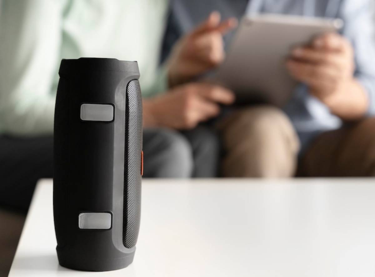  A black portable speaker sits on a white table, with two people discussing and using a tablet in the blurred background.