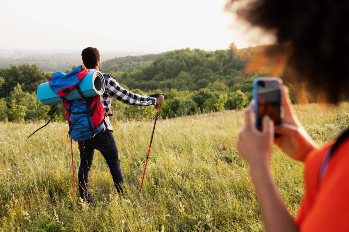 A hiker with a blue and red backpack stands on a hillside, gazing at the scenic view, while a friend captures the moment on their phone. 