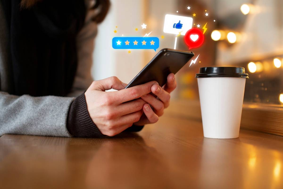 A person holds a smartphone displaying social media reactions, with a coffee cup on a table in a cozy cafe setting.