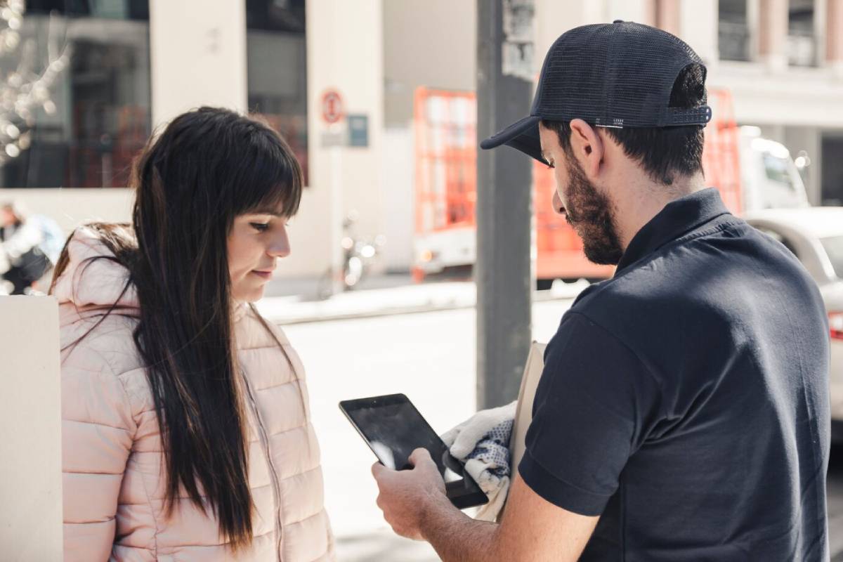A woman in a light pink jacket stands beside a man in a black cap, who holds a tablet, engaged in conversation on a city street.