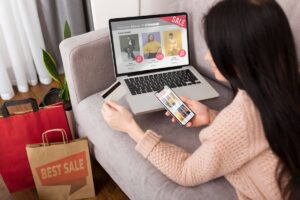 A woman sits on a couch shopping online using a laptop and smartphone, surrounded by shopping bags.