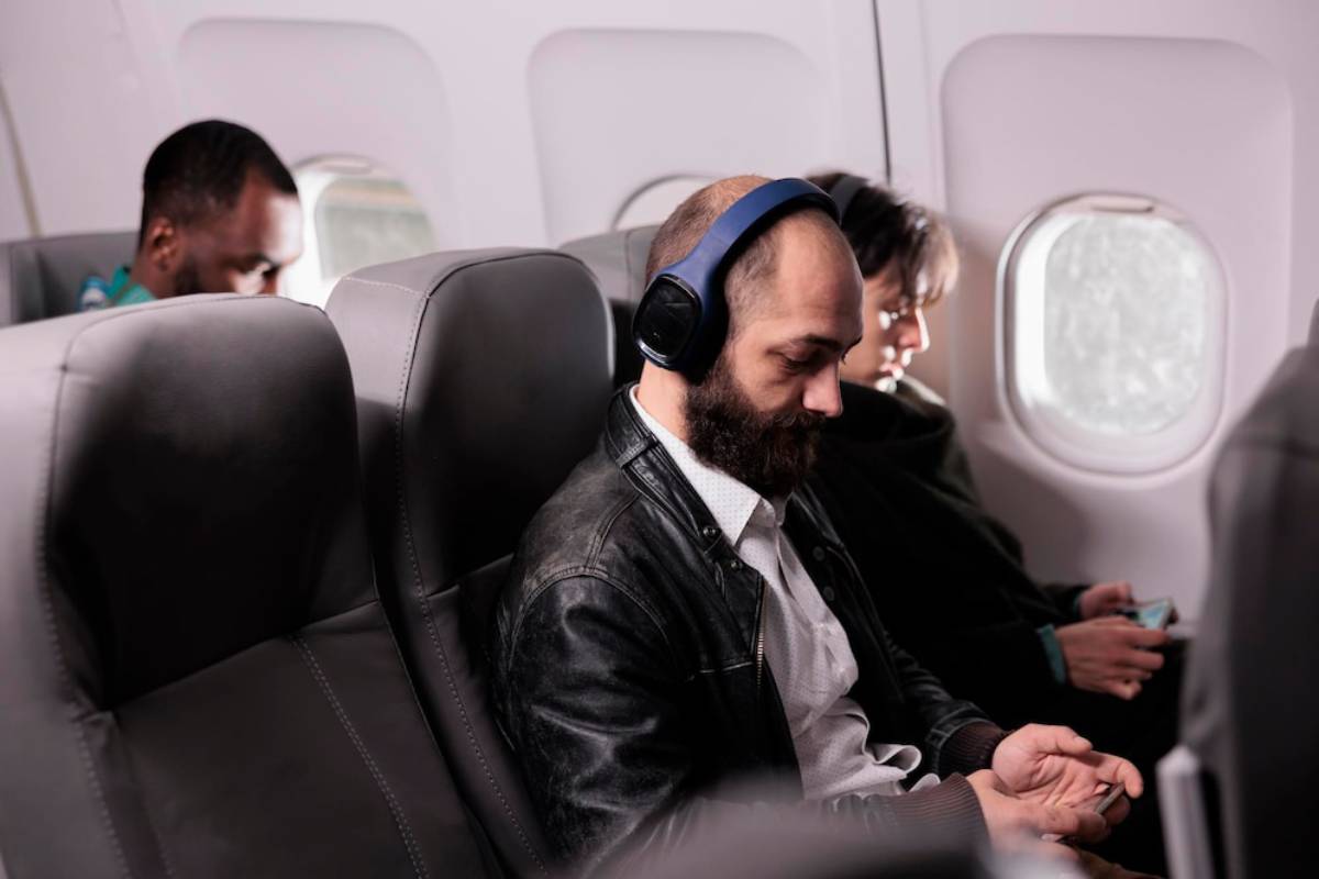 Passengers sitting close together in an airplane, one wearing headphones, another holding a device, with cloudy landscape visible through the window.
