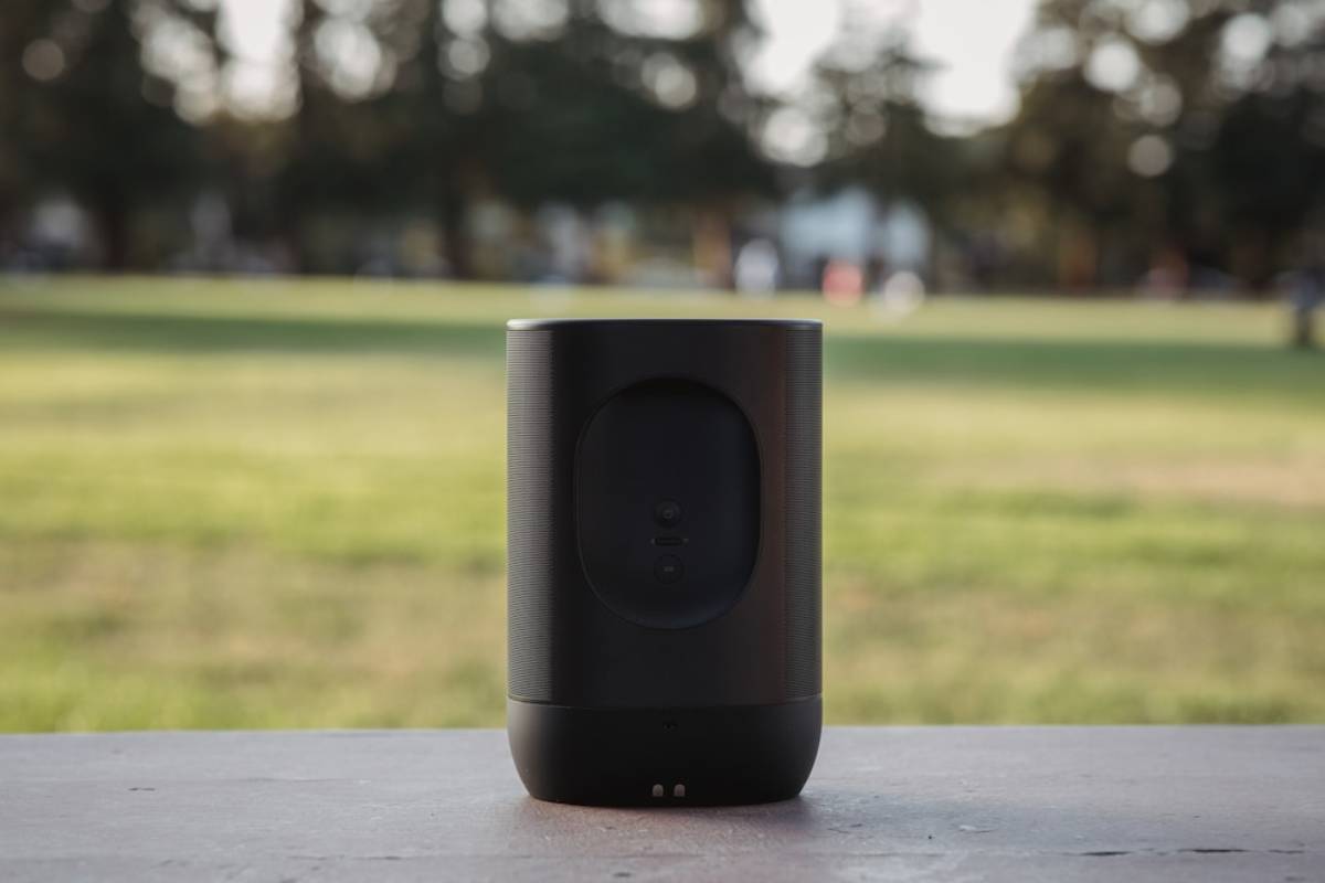 A sleek black speaker sits on a wooden surface, set against a blurred green park background. The speaker features buttons for operation.