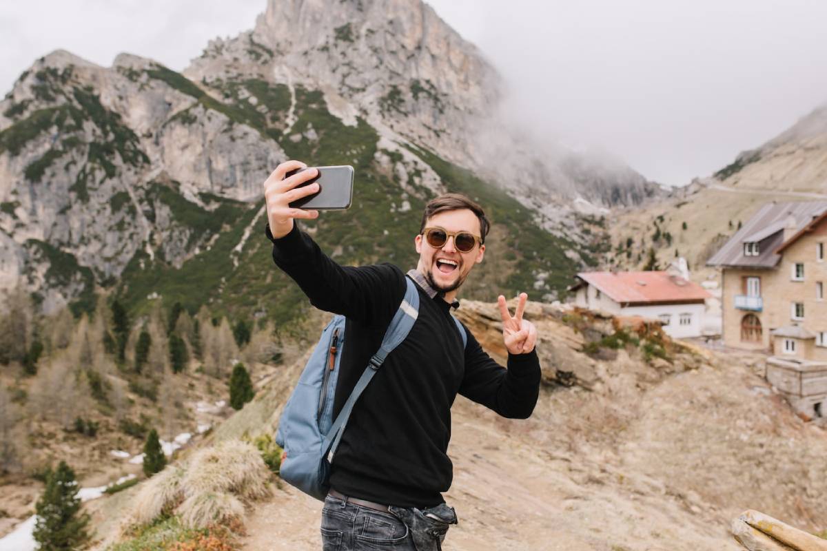 A person with a backpack takes a selfie in a scenic mountainous landscape, showcasing the lush green hills and cloudy skies. 