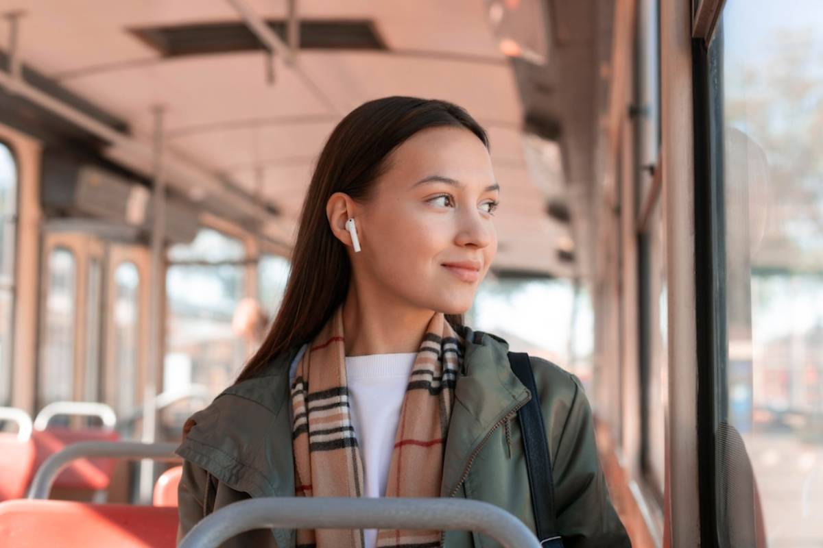 A woman wearing AirPods stands inside a bus, gazing out the window, dressed in a green jacket and a plaid scarf.