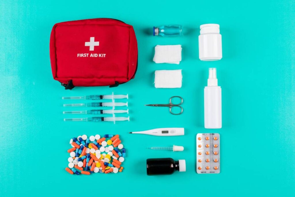 A flat lay of a red first aid kit, syringes, medications, bandages, scissors, and a thermometer, all on a teal background.