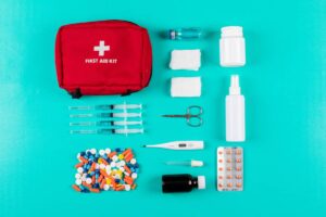 A flat lay of a red first aid kit, syringes, medications, bandages, scissors, and a thermometer, all on a teal background.