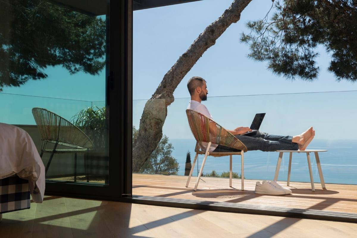A person sits on a balcony, working on a laptop, overlooking a scenic ocean view and surrounded by greenery and modern decor. 