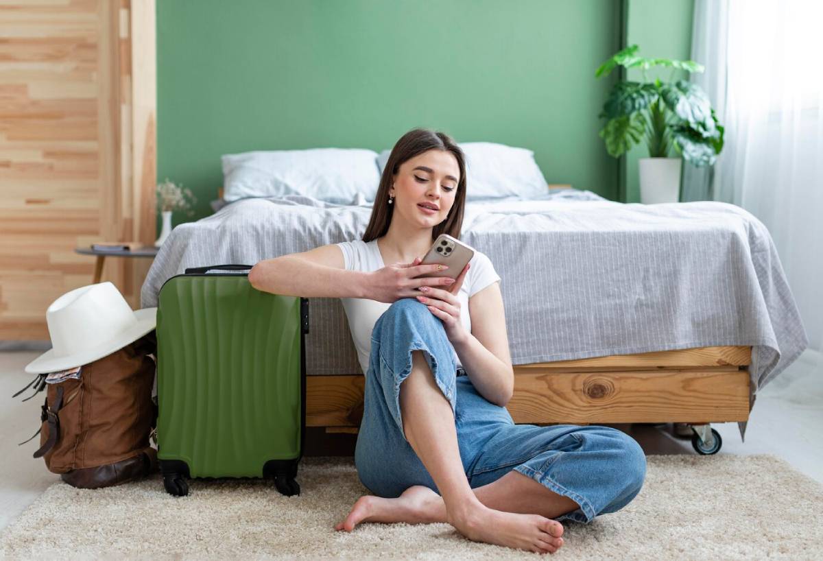 A person sits on the floor next to a green suitcase, wearing casual clothes, looking at their phone in a cozy, green-themed bedroom.