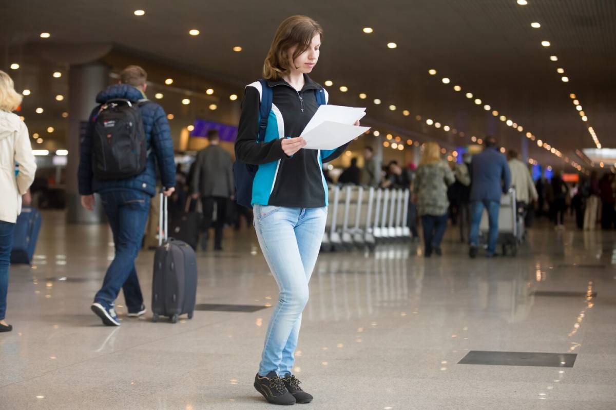 A person stands in an airport terminal, holding documents, surrounded by other travelers and luggage on a busy day. 