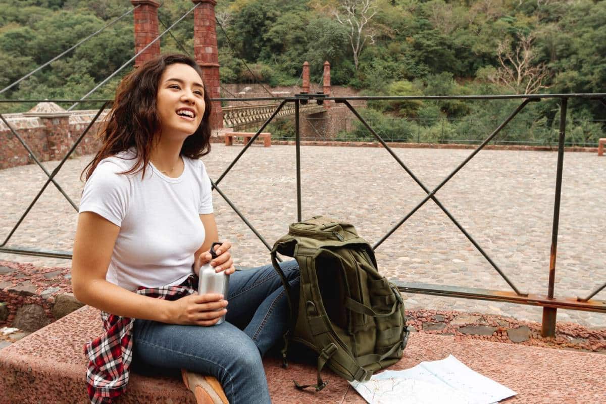 A person sits on a stone bench, holding a water bottle, with a backpack and a map beside them, surrounded by greenery and a pathway.