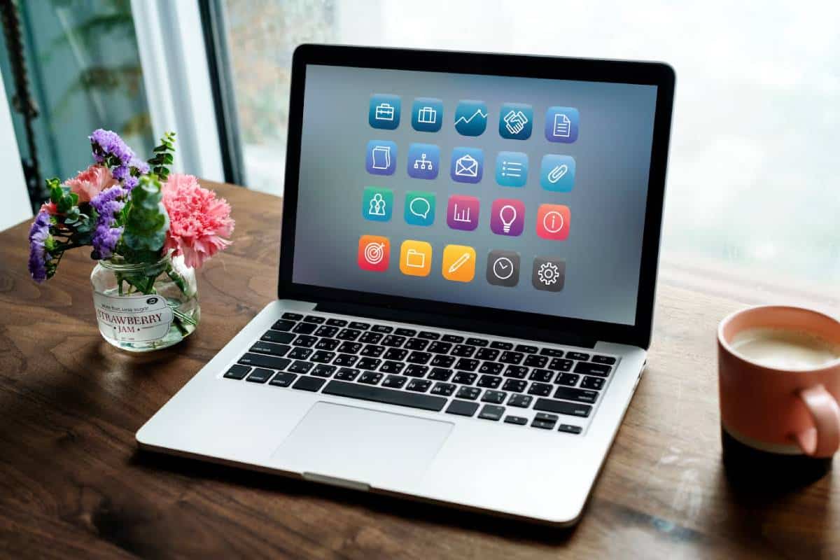 A laptop on a wooden table displays colorful app icons, next to a cup of coffee and a small vase of flowers.