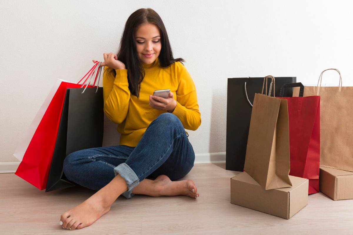 A woman in a yellow sweater sits on the floor with shopping bags around her, engrossed in her phone. 