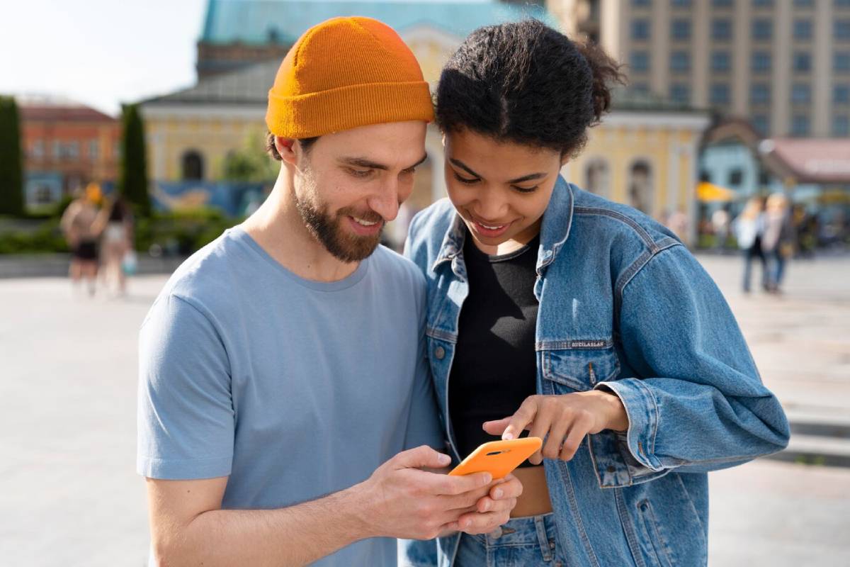 A man in a blue shirt and a woman in a denim jacket share a moment, looking at a smartphone outdoors in a lively urban setting.