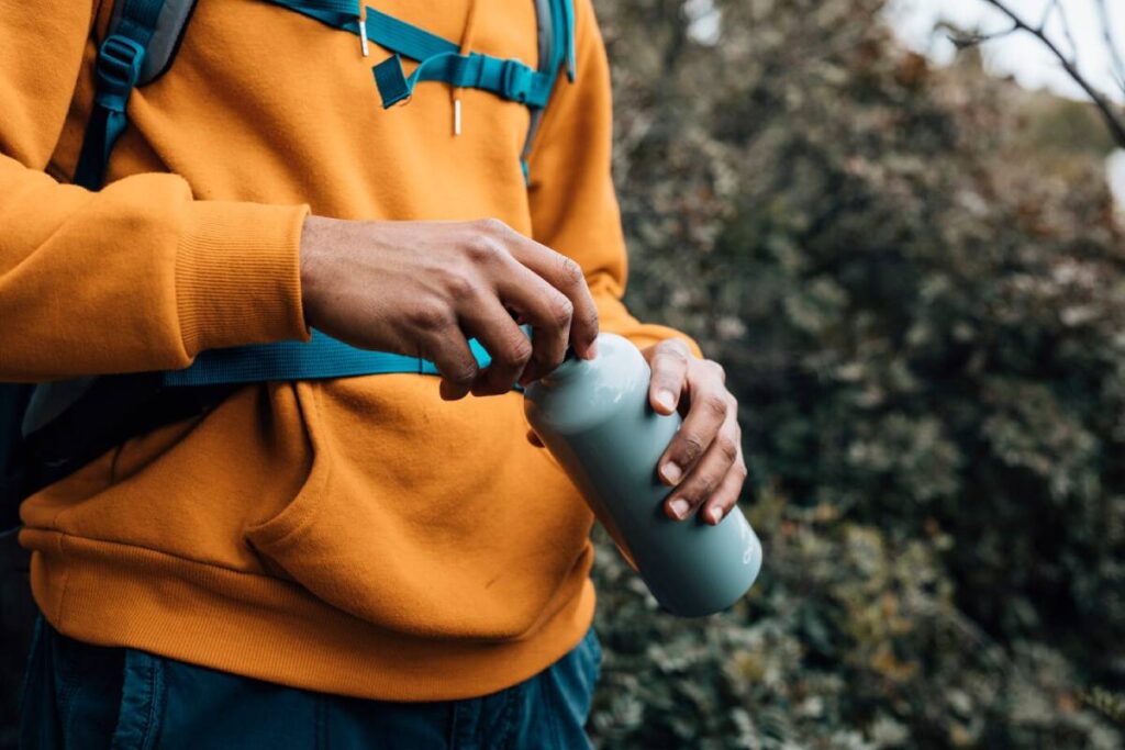 A person in an orange sweatshirt opens a gray water bottle while standing outdoors, surrounded by greenery.