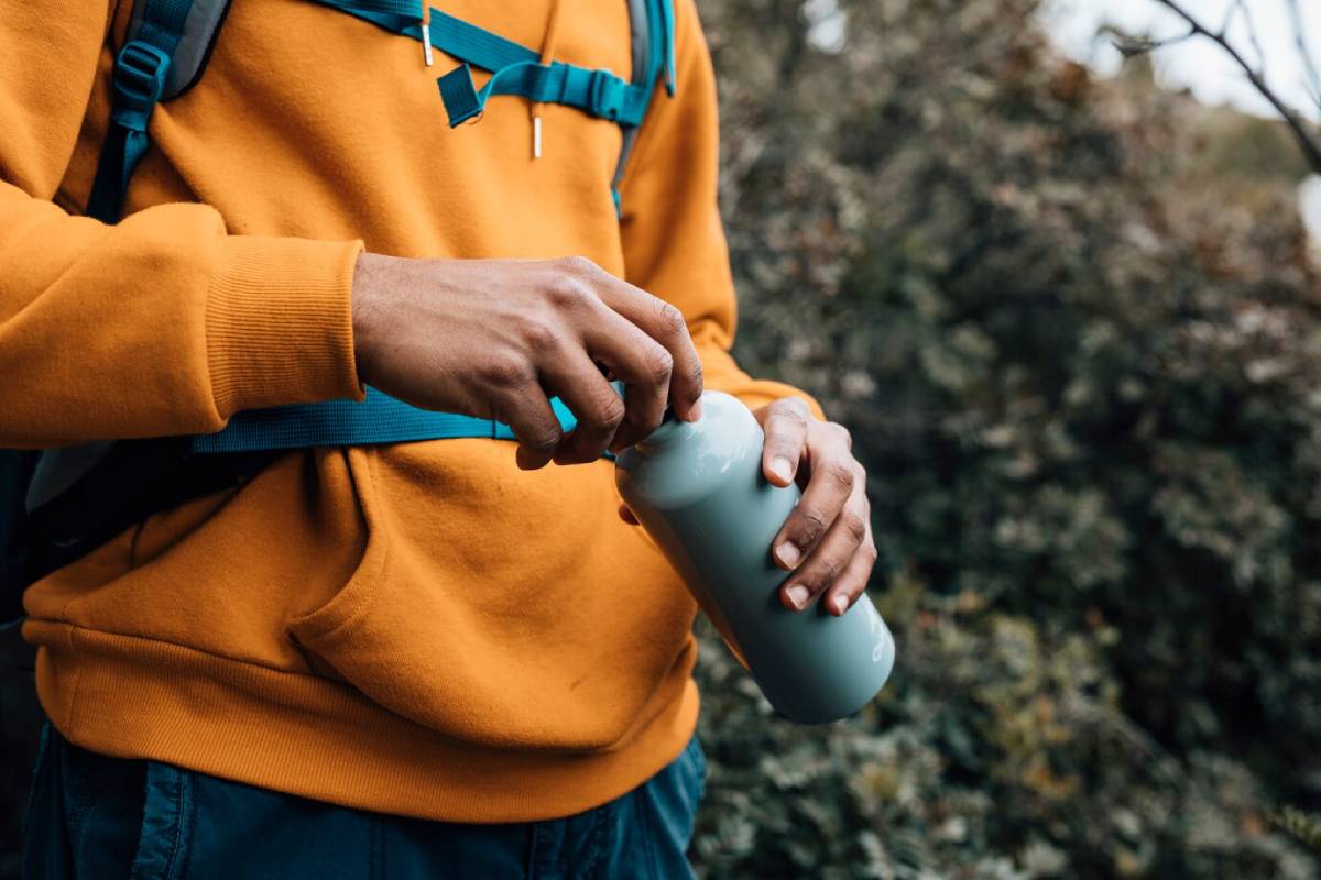 A person in an orange sweatshirt opens a gray water bottle while standing outdoors, surrounded by greenery.