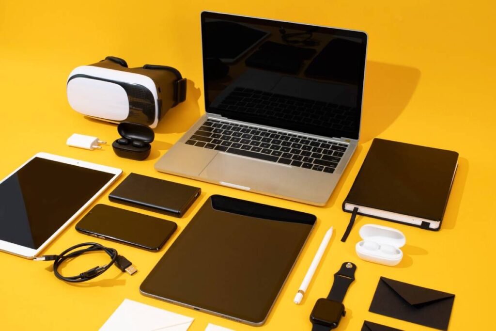 A flat lay of modern tech gadgets: a laptop, tablet, smartphones, VR headset, smartwatch, earbuds, notebooks, and charging accessories on a yellow background.