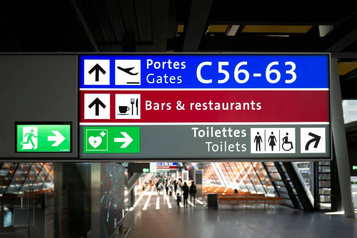 A directional airport sign indicating gates C56-63, bars/restaurants, toilets, and emergency services with corresponding icons. 
