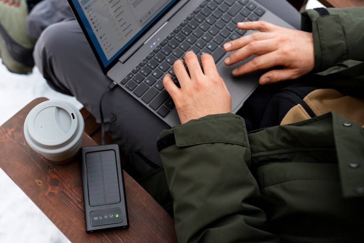A person types on a laptop while sitting outdoors, with a to-go coffee cup and a solar charger on a wooden table.