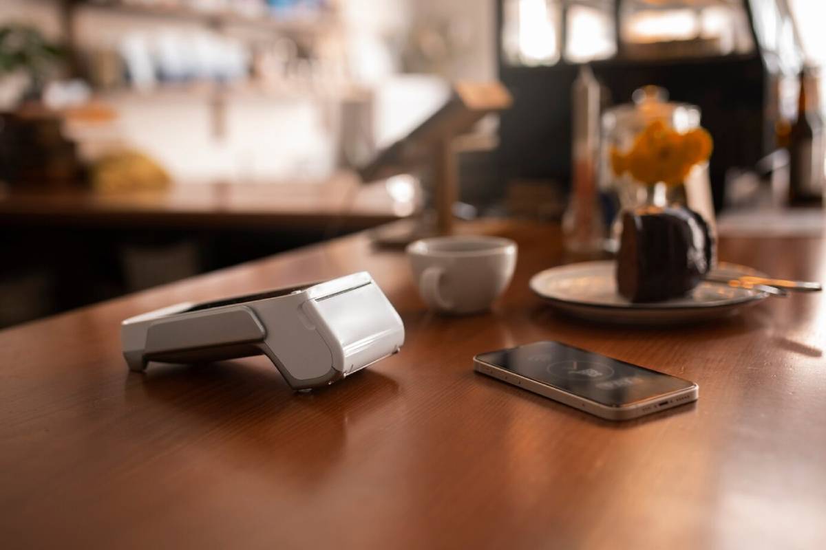 A smartphone and a white payment device sit on a wooden table beside a coffee cup and a plate of chocolate cake. 