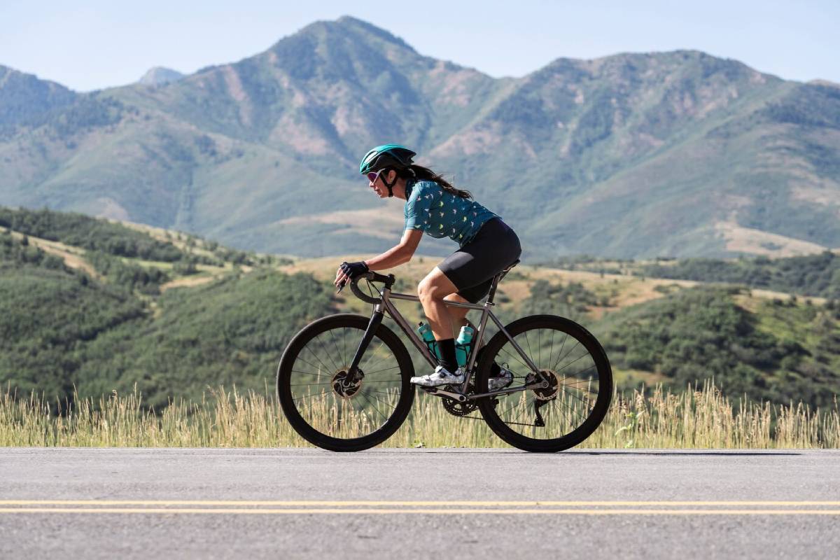 A cyclist rides along a road, with lush green hills and mountains in the background under a clear blue sky. 