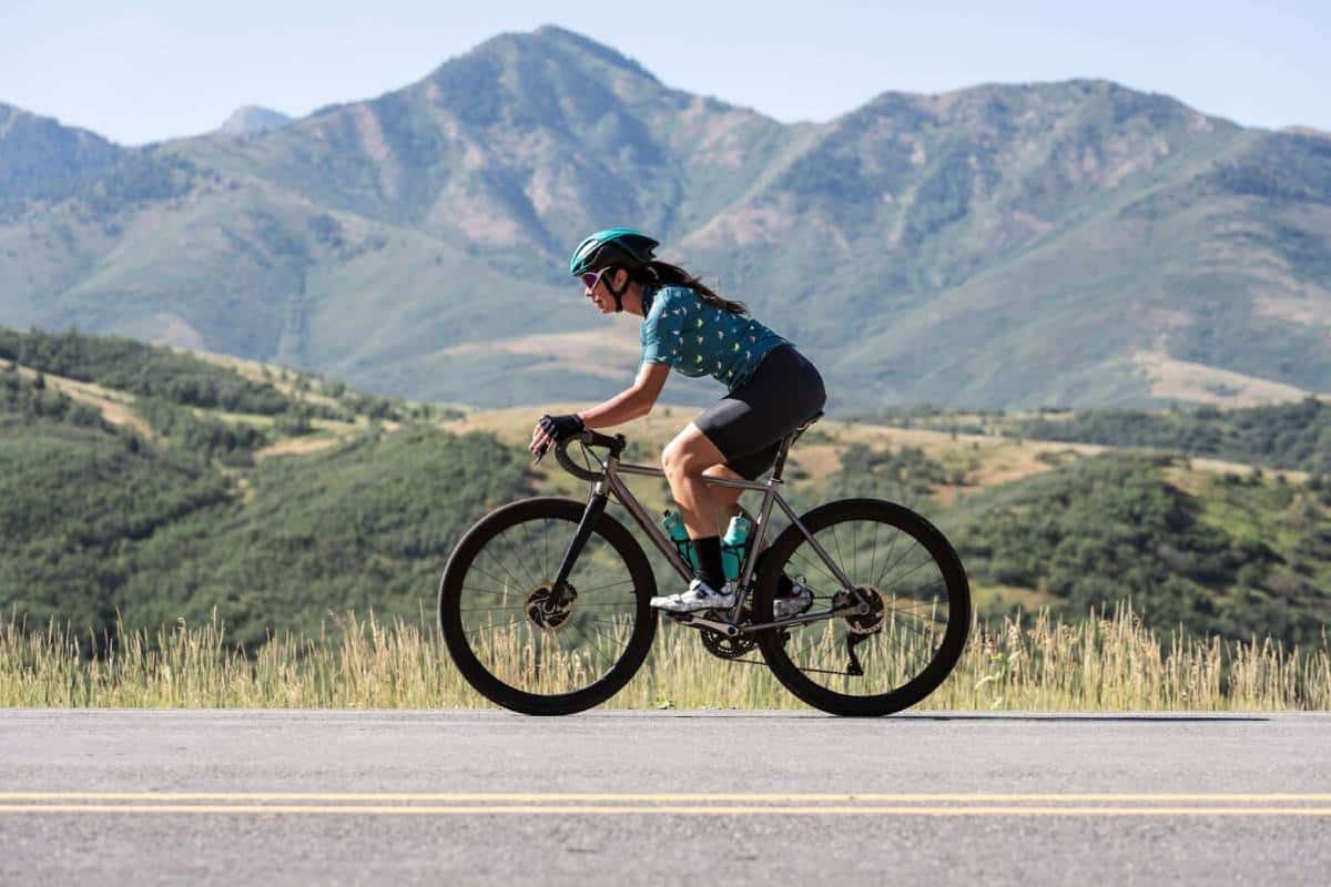 A cyclist rides along a road, with lush green hills and mountains in the background under a clear blue sky.