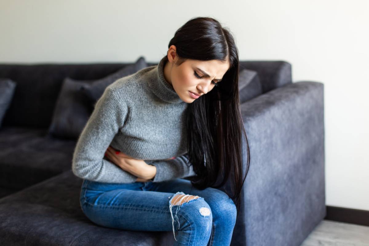 A person sits on a couch, wearing a gray sweater and jeans, holding their stomach, appearing to be in discomfort.
