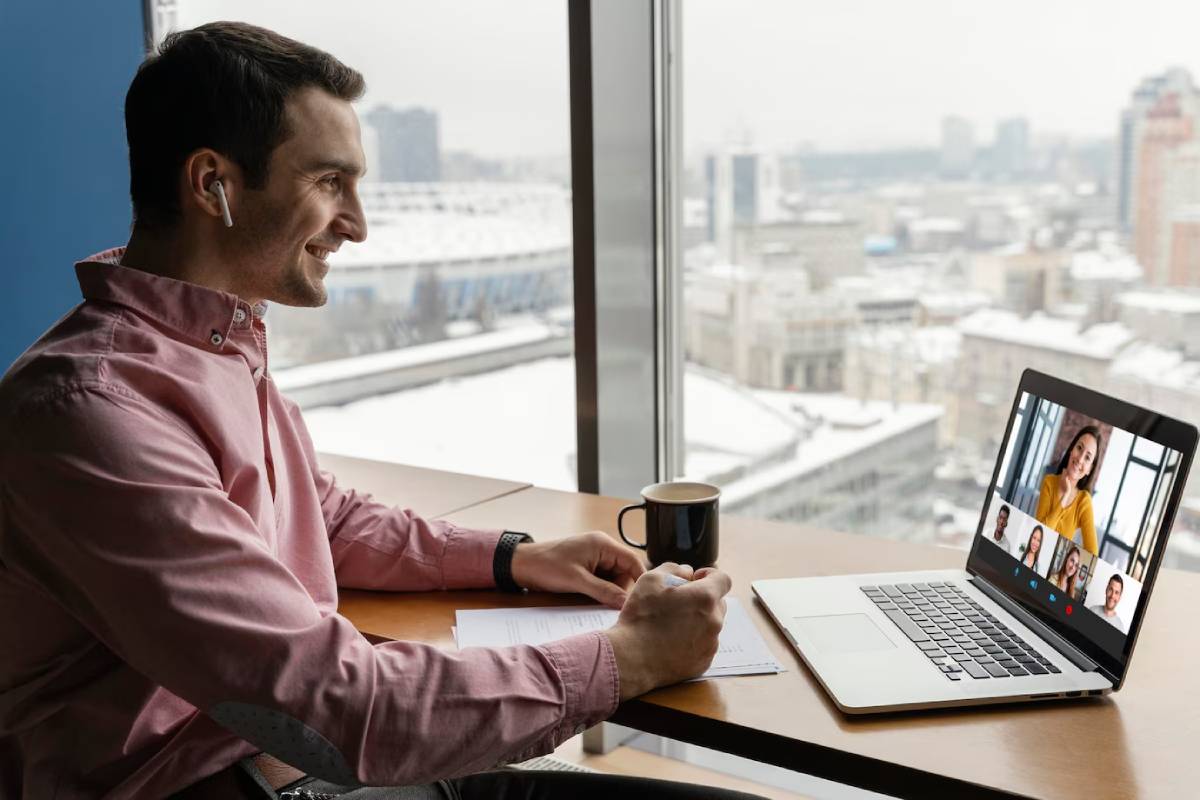 A person in a pink shirt uses a laptop for a video call while holding a coffee mug, with a city skyline visible outside. 