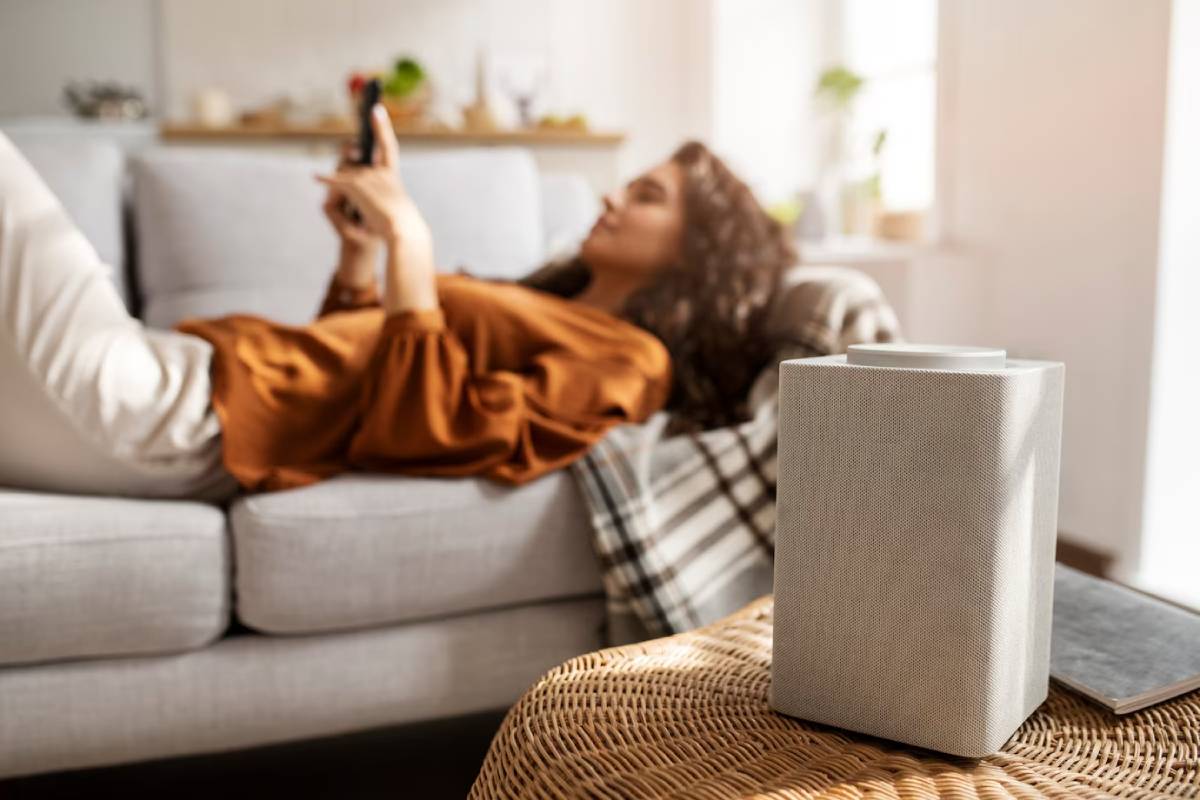 A cozy living room scene featuring a modern speaker on a woven table, with a person relaxing on a couch in the background.