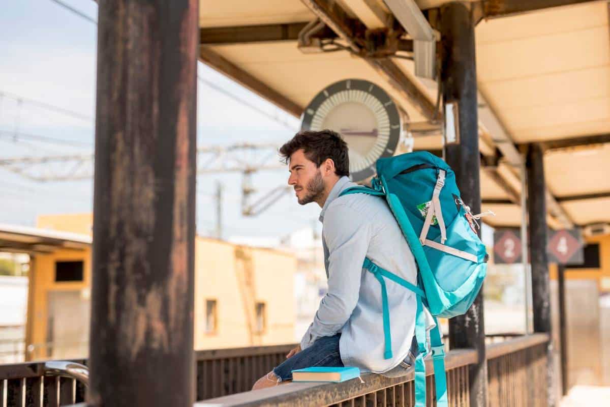 A person sits on a platform with a large teal backpack, gazing at a nearby clock, surrounded by an urban train station.