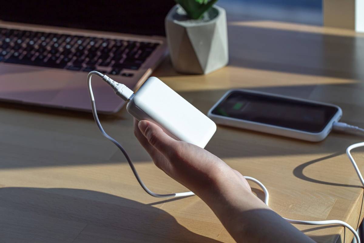 A hand holds a white charger, connected to a laptop and a phone on a wooden table, with sunlight illuminating the scene.
