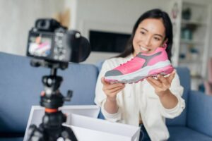 A person holds a vibrant pink sneaker while filming a review in a cozy living room setting. A camera is positioned to capture the shoe.