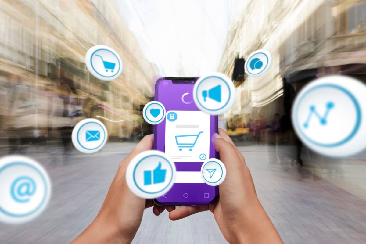  A person holds a smartphone displaying a shopping cart icon, surrounded by social media and communication symbols, in a blurred urban setting.