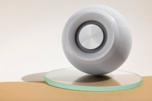 A sleek, modern speaker with a circular design rests on a glass surface, showcasing its minimalist style against a neutral backdrop.