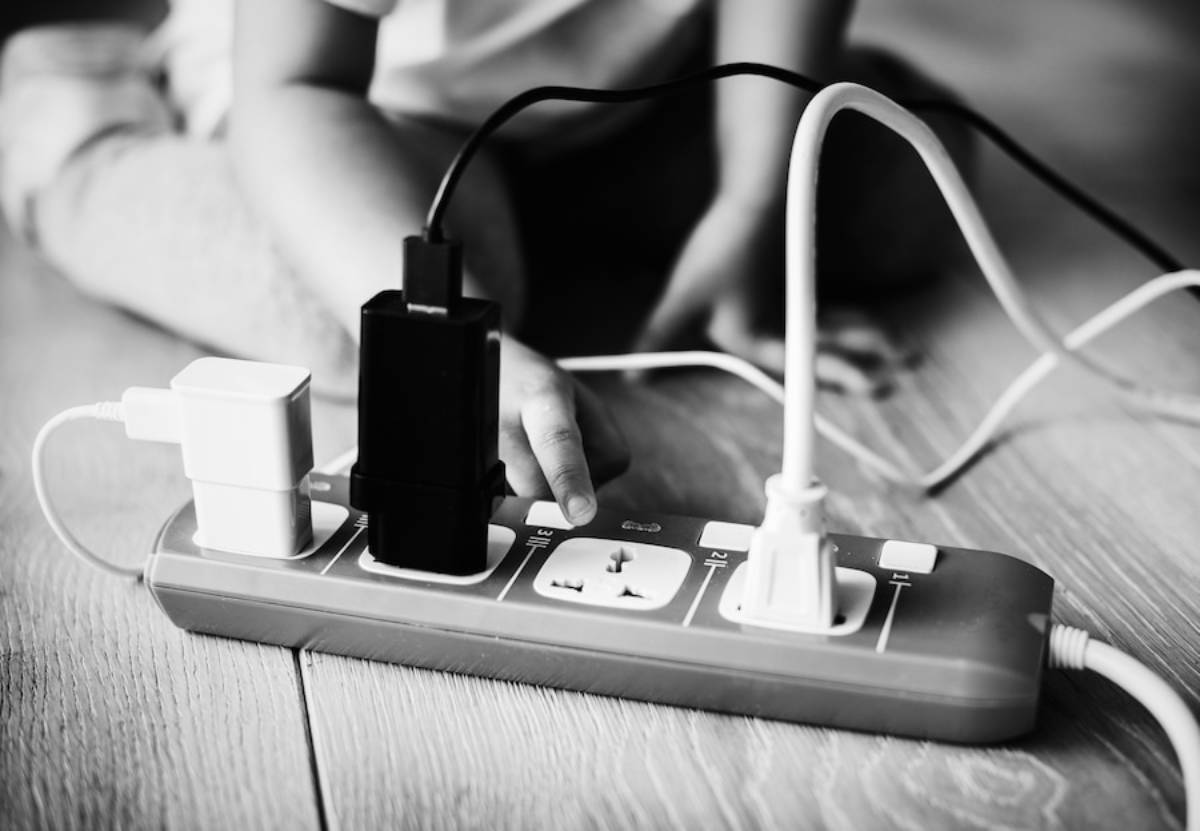 A child's hand is reaching for a power strip with multiple plugs, including a white and a black charger, on a wooden floor.