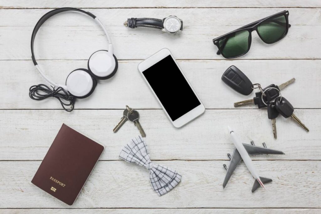 Flat lay of travel essentials: headphones, smartphone, sunglasses, keys, watch, bow tie, passport, and a toy airplane on wooden surface.