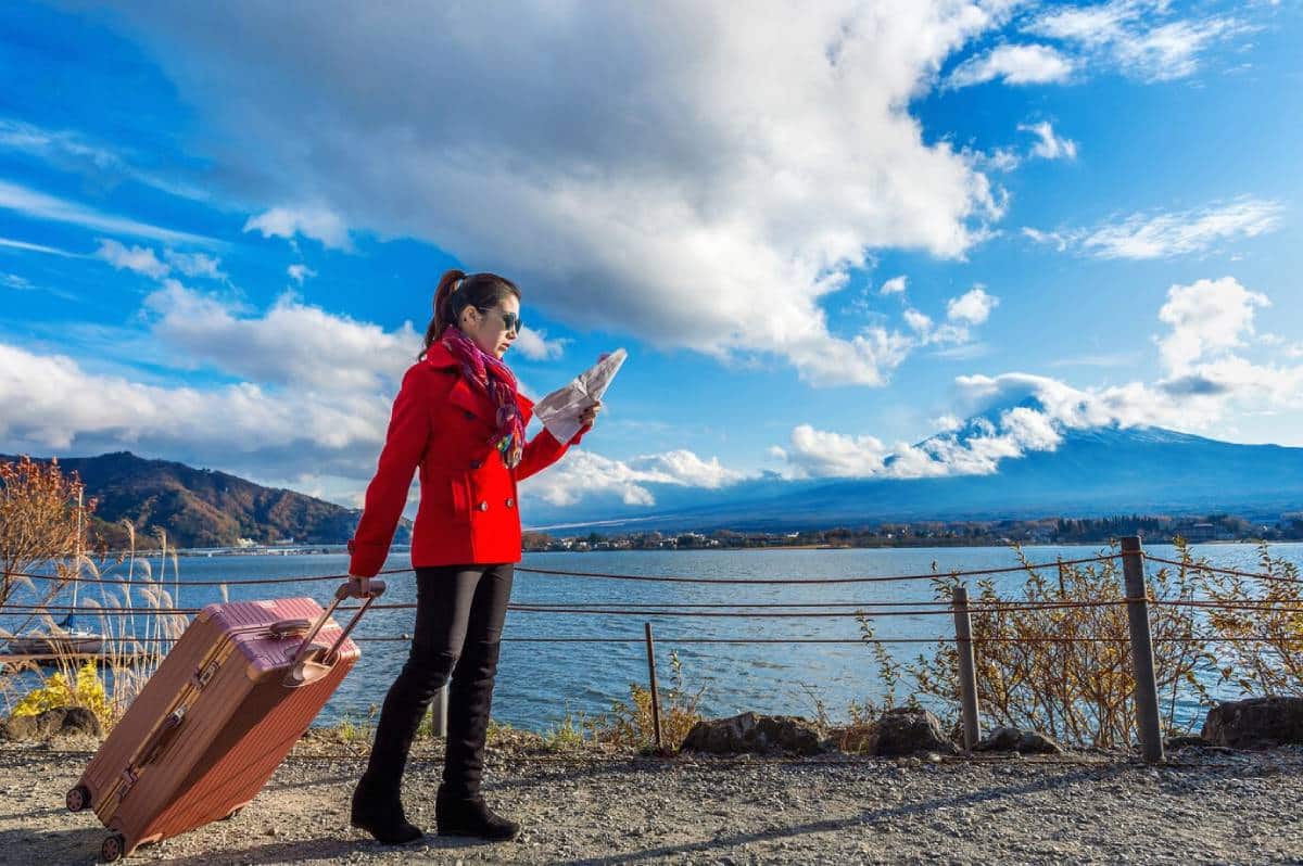A woman in a red jacket stands by a lake with a pink suitcase, holding a map, with mountains and blue skies in the background.